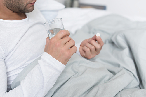 Cropped image of Sick man holding pill and glass of water in hands lying in a bed