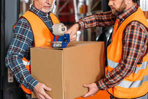 cropped view of male workers in helmets packing cardboard box with scotch tape