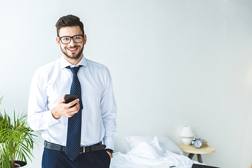 smiling businessman in eyeglasses using smartphone in bedroom