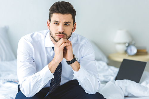 handsome thoughtful businessman in white shirt and tie sitting on bed