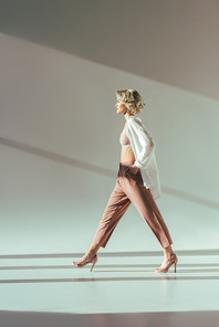 side view of stylish blonde woman in pink bra, shirt and pants walking in studio on grey