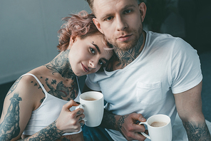 young tattooed couple looking at camera and drinking coffee in bed at morning
