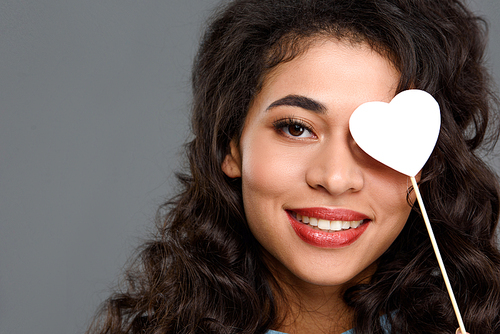 close-up portrait of smiling young woman covering eye with heart on stick isolated on grey