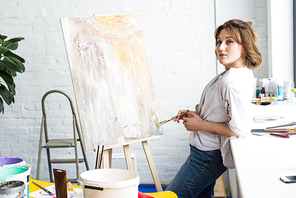 Young artistic girl standing by easel with brush in light studio