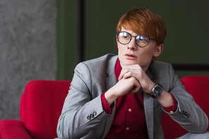 Male fashion model in eyeglasses dressed in suit sitting on red couch