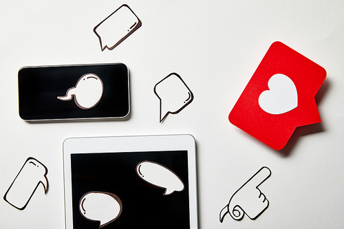 digital tablet and smartphone with paper thought and speech bubbles, paper heart, pointing card on white surface