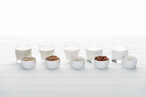 glasses with coconut, chickpea, oat, drick and almond milk on white wooden surface with ingredients in bowls isolated on white