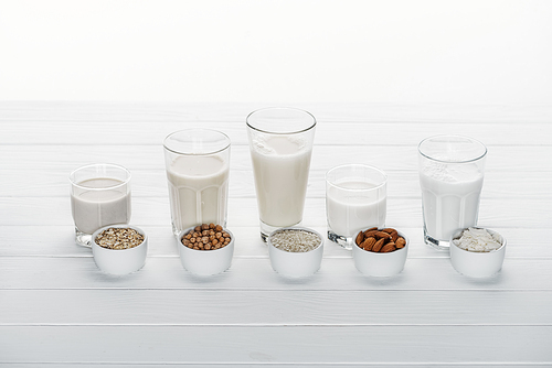 glasses with coconut, chickpea, oat, drick and almond milk on white wooden table with ingredients in bowls isolated on white