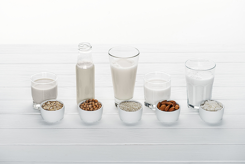 glasses and bottle with coconut, chickpea, oat, drick and almond milk on white wooden surface with ingredients in bowls isolated on white