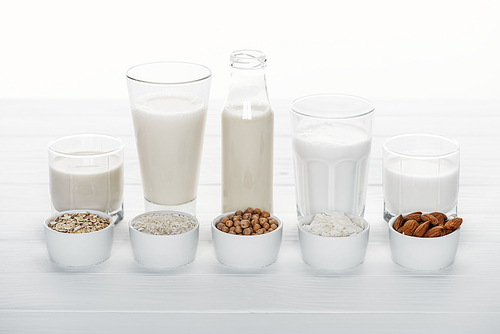 glasses and bottle with coconut, chickpea, oat, drick and almond milk on white wooden table with ingredients in bowls isolated on white