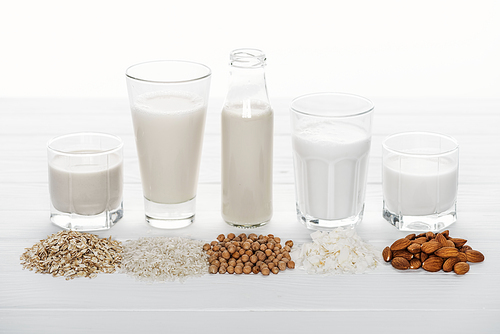glasses and bottle with coconut, chickpea, oat, drick and almond milk on white wooden surface with ingredients isolated on white