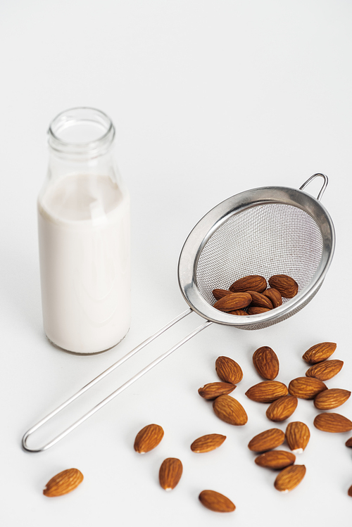 vegan almond milk in bottle near scattered almonds and sieve on grey background