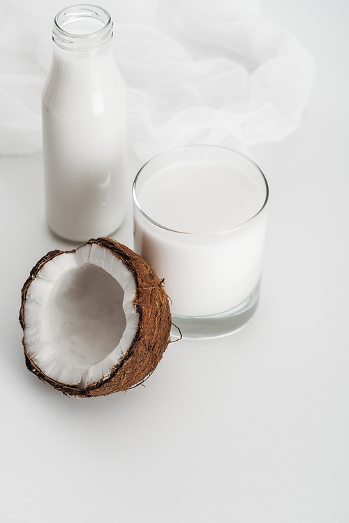 organic vegan coconut milk in glass and bottle near coconut half and cheesecloth on grey background