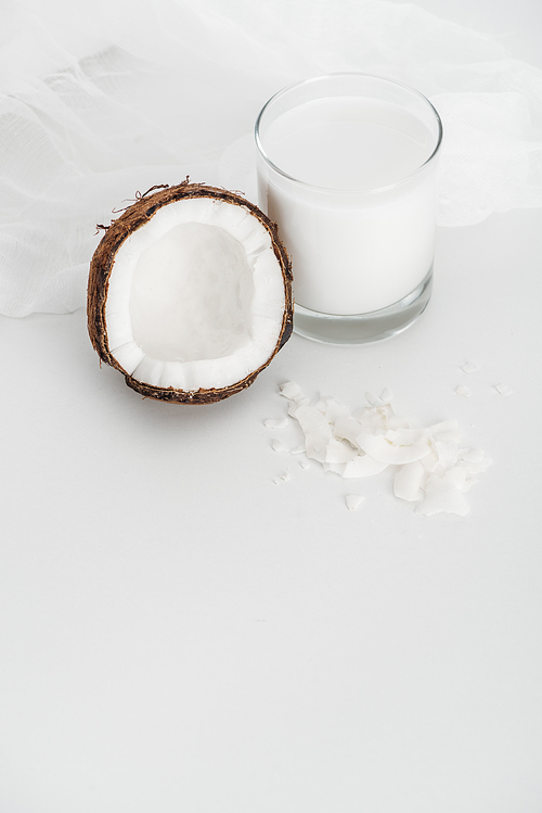 organic vegan coconut milk in glass near coconut half, chips and cheesecloth on grey background