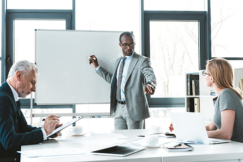 young african american businessman writing on whiteboard and looking at colleagues at business meeting