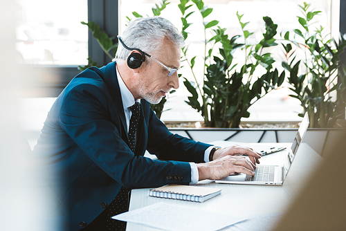 side view of senior businessman in eyeglasses and headset using laptop in office