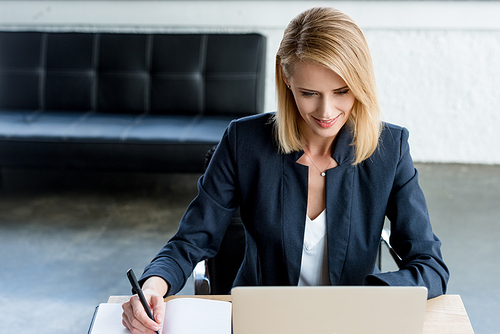 high angle view of smiling businesswoman taking notes and working with laptop in office