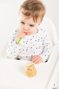 high angle view of baby boy eating puree from jar and sitting in highchair isolated on white