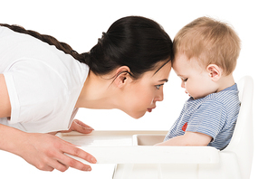 side view of mother touching forehead of little son sitting in highchair isolated on white