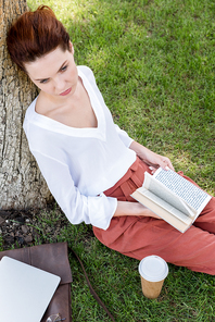 high angle view of beautiful young woman with book leaning back on tree trunk in park