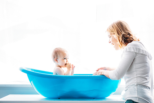 side view of smiling mother washing her adorable little child in plastic baby bathtub at home