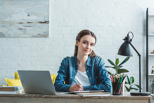 teenage girl taking notes and smiling at camera while studying with laptop at home