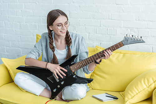 smiling teen girl playing electric guitar while sitting on yellow sofa with textbook