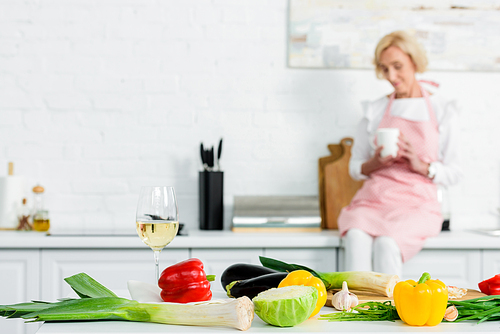 beautiful senior woman sitting on kitchen counter with cup of tea with vegetables on foreground
