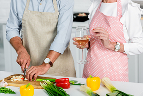 cropped image of senior husband cutting vegetables and wife holding glass of wine at kitchen