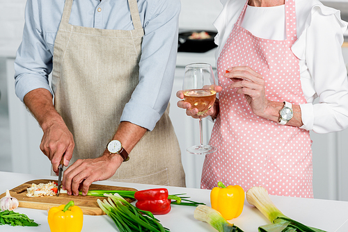 cropped image of senior husband cutting vegetables and wife holding glass of wine at kitchen