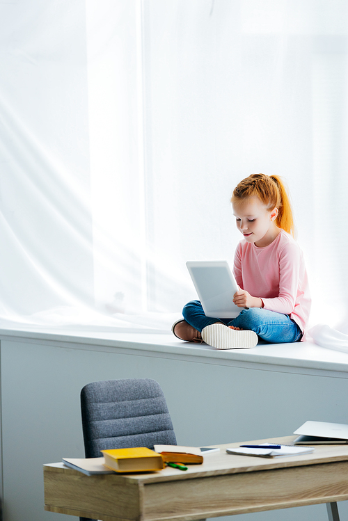 adorable child using digital tablet while sitting on windowsill