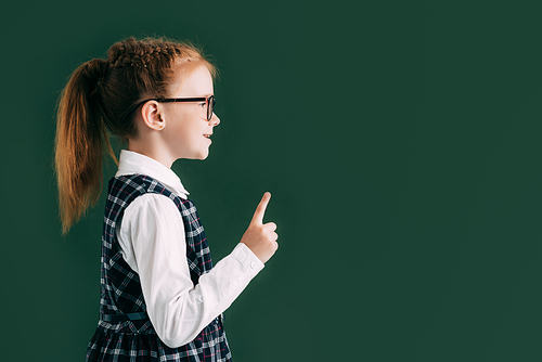 side view of little smiling schoolgirl in eyeglasses pointing with finger while standing near blackboard