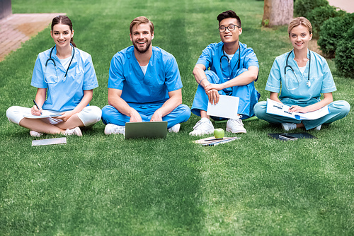 smiling multicultural medical students sitting on grass and
