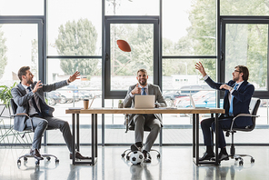 smiling young businessmen using laptops and playing with soccer and rugby balls in office