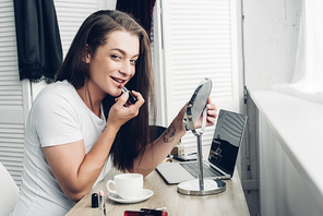 smiling young transgender man with round makeup mirror applying lipstick while 