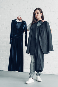 young transgender man holding male suit and dress in front of white brick wall