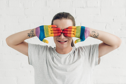 smiling transgender man covering eyes with hands painted in colors of pride flag in front of white brick wall