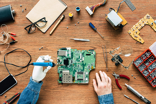cropped image of electronic engineer with robotic hand fixing motherboard by soldering iron at table surrounded by tools