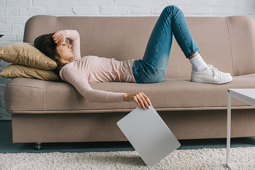 side view of young woman with headache lying on sofa and holding laptop