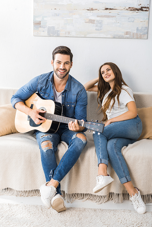 smiling young man playing on acoustic guitar while his girlfriend sitting near on couch at home