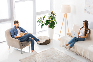 high angle view of woman using laptop on sofa while her boyfriend reading book on armchair at home