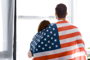 rear view of couple covered with american flag looking out window at home
