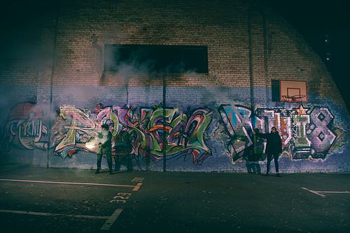 people holding smoke bombs and standing against wall with graffiti at night