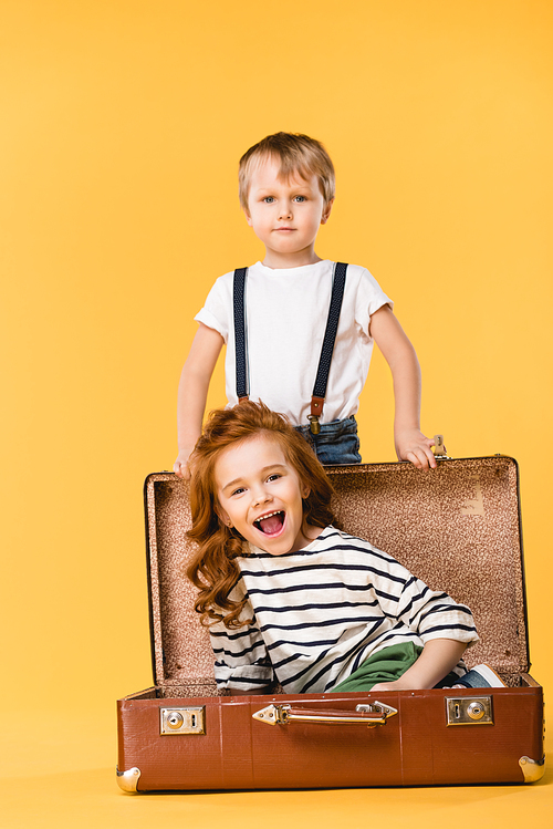 happy kid sitting in suitcase with boy standing near by isolated on yellow