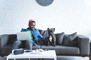 African american man working on laptop and hugging French bulldog by table with modern gadgets