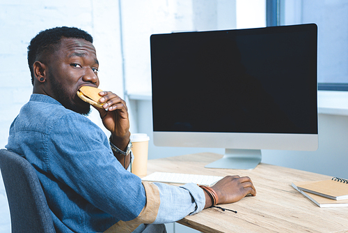 African american man working by computer and eating hamburger