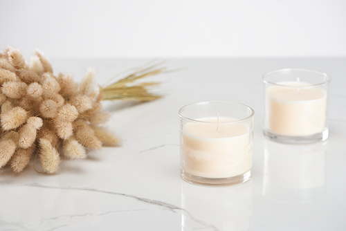 selective focus of fluffy bunny tail grass and white candles in glass on marble white surface