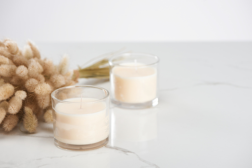 selective focus of fluffy bunny tail grass and white candles in glass on marble white surface