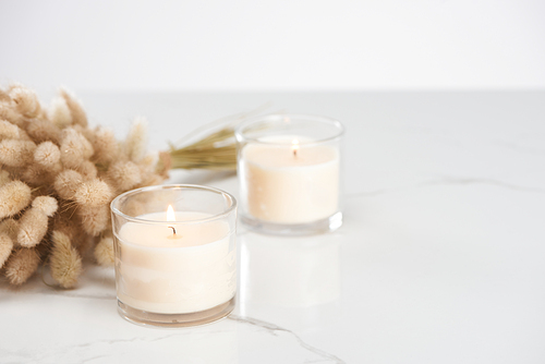 selective focus of fluffy bunny tail grass and burning white candles in glass glowing on marble white surface