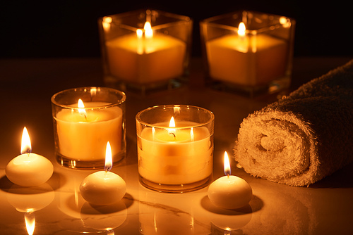 selective focus of burning candles in glass glowing in dark near rolled towel on marble surface